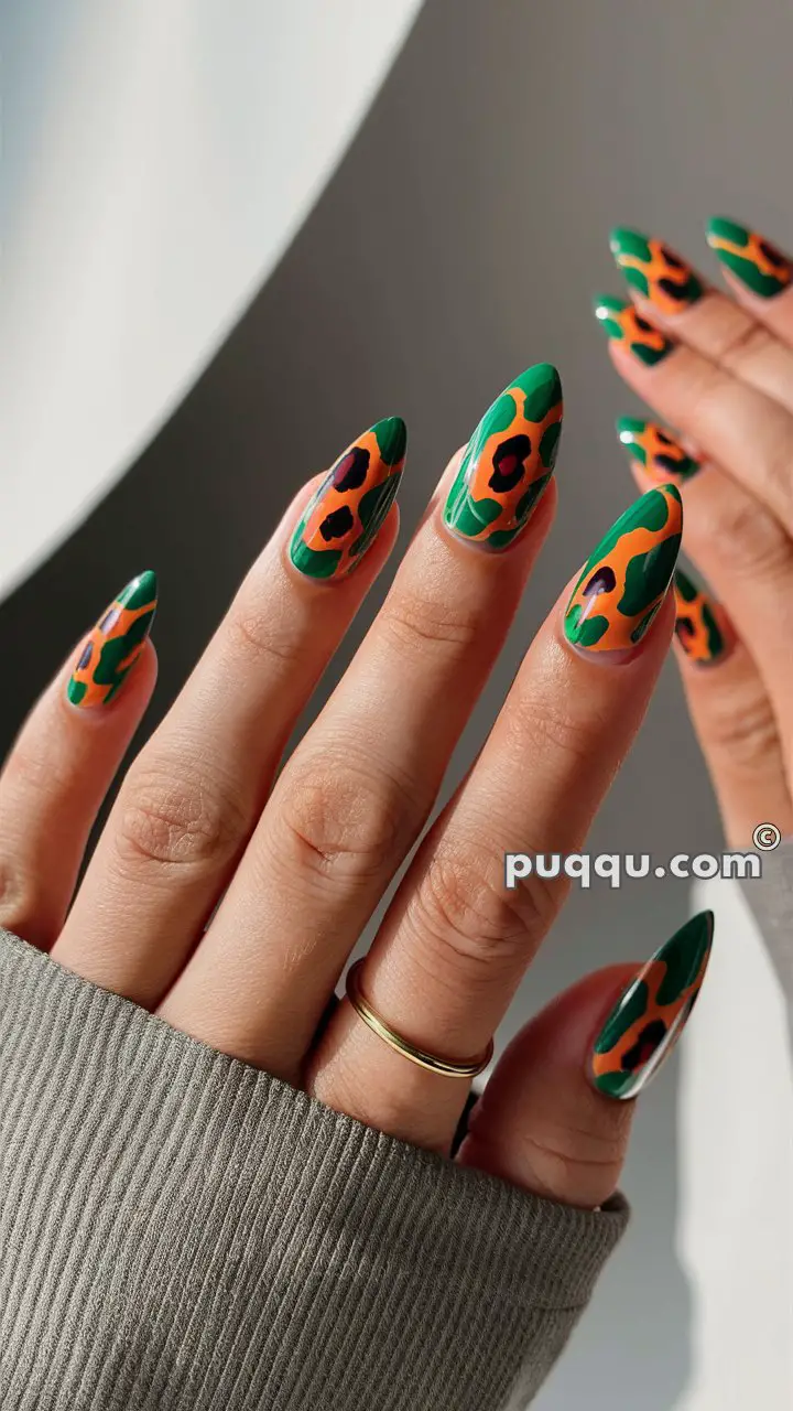 Almond-shaped nails with an abstract floral design in green, orange, and black, hand wearing a beige sweater and gold ring.