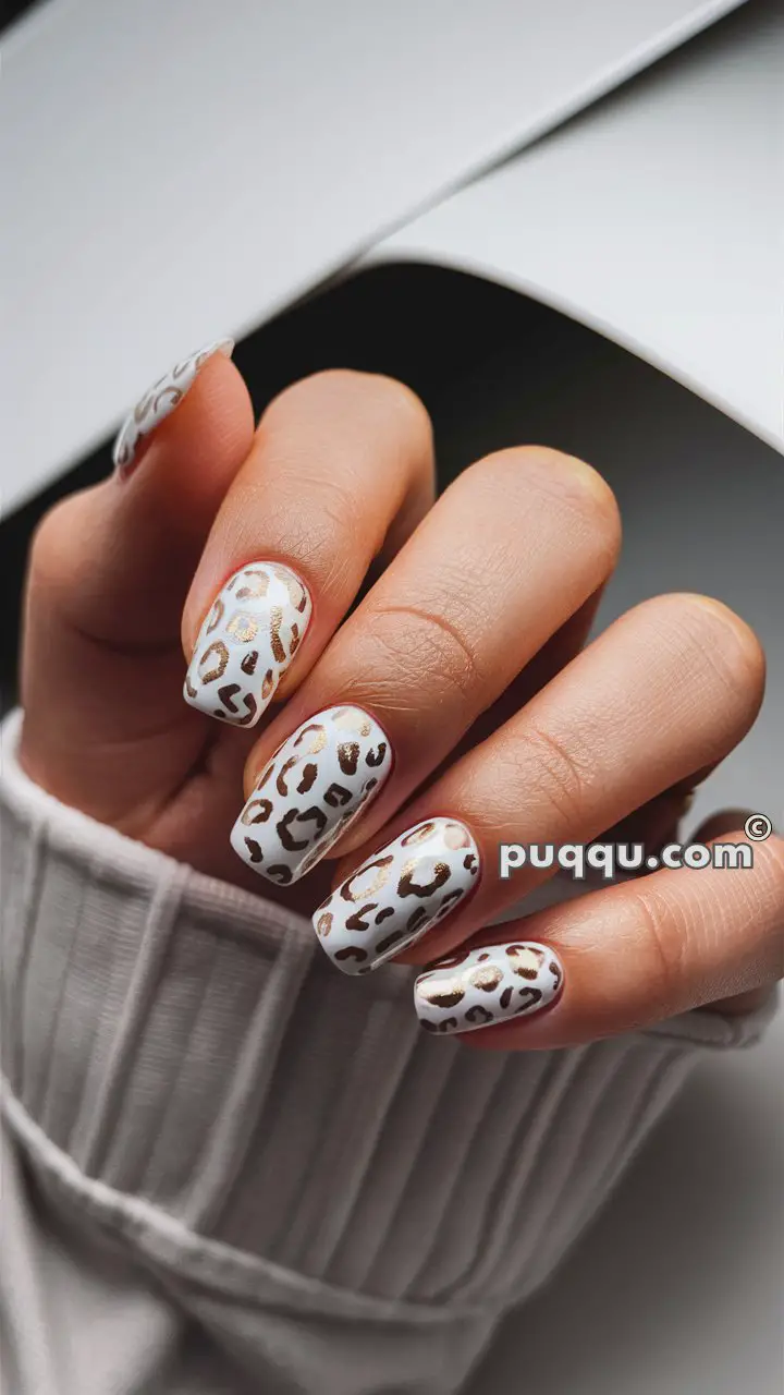 A hand with long, white nails featuring a gold and brown leopard print design, wearing a knitted white sweater.