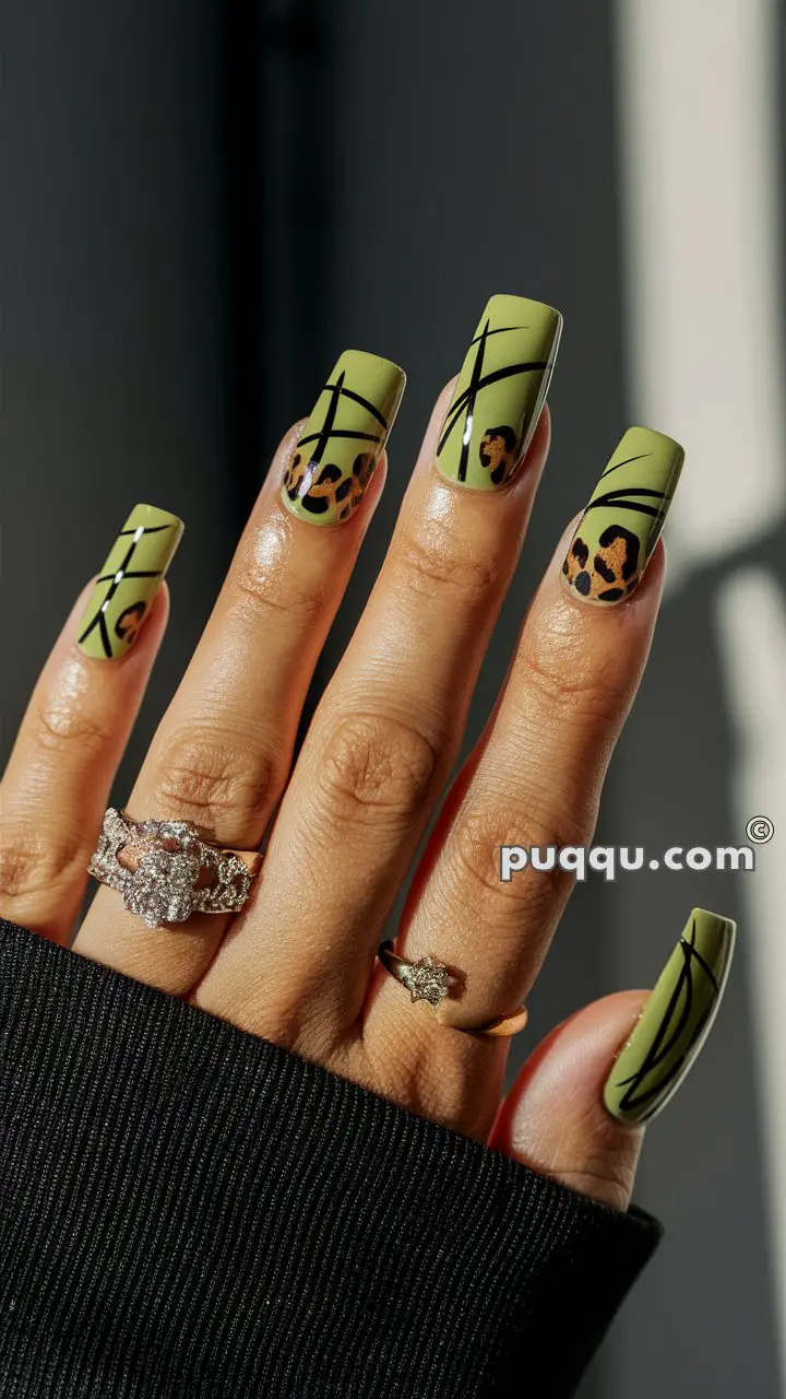 Close-up of a hand with long, green nails featuring black diagonal lines and leopard print accents, adorned with multiple rings.