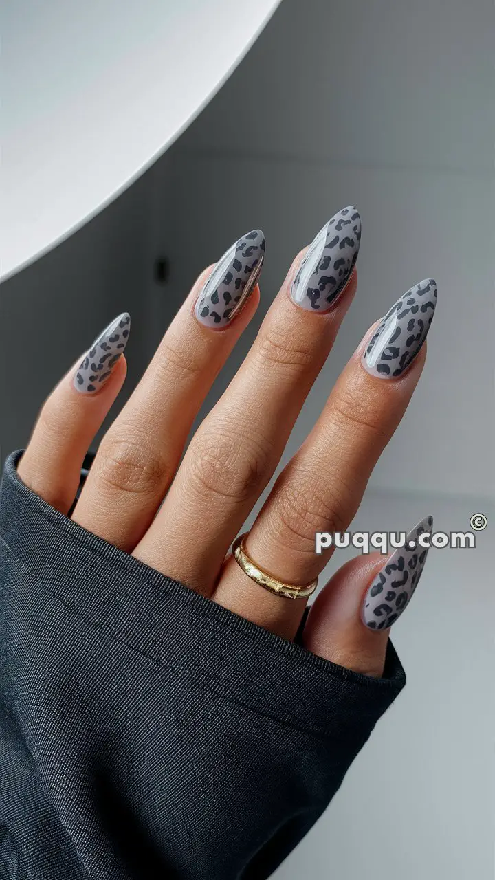 Hand with long nails featuring gray and black leopard print design, wearing a gold ring and dark sleeve.