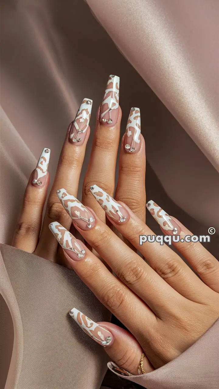 Hands with long, coffin-shaped nails featuring white and beige animal print design and small rhinestones.