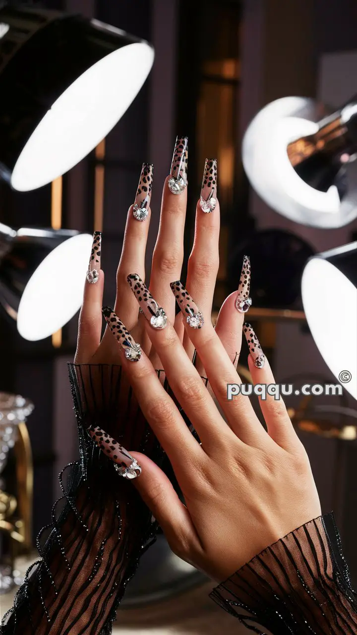 Hands with long, pointed nails featuring a black and clear spotted pattern and adorned with large rhinestones.