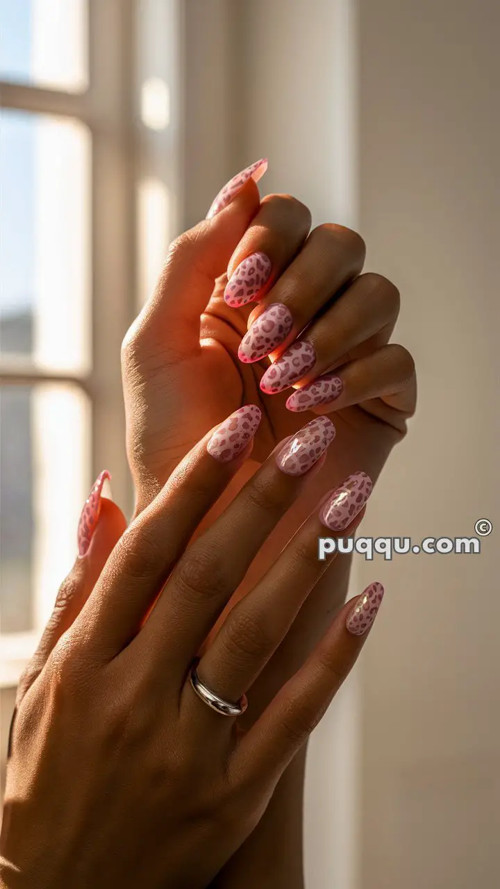 Hands with long, oval-shaped nails painted with a pink and purple leopard print design, wearing a silver ring, in front of a window with natural light.