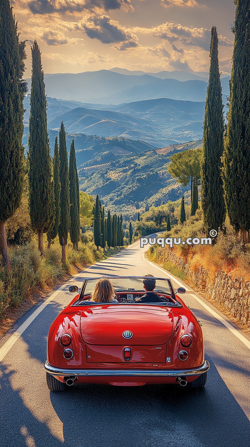 Two people driving a red convertible car on a scenic road lined with cypress trees and overlooking a mountainous landscape at sunset.