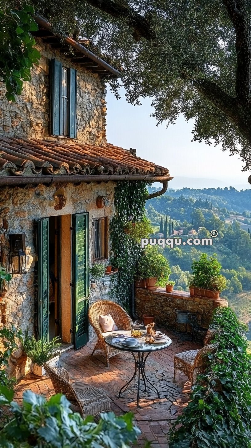 Stone house with green shutters, a terracotta tile roof, and a charming terrace featuring wicker chairs and a small round table set for a meal, surrounded by lush greenery and a scenic hillside view.