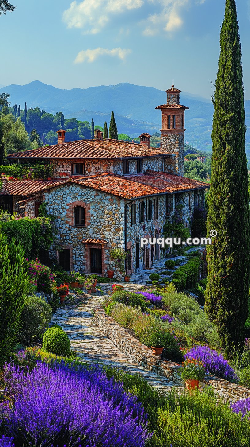 Stone villa with red tile roof, tall cypress trees, and vibrant purple flowers in a lush garden setting against a backdrop of rolling hills and mountains.