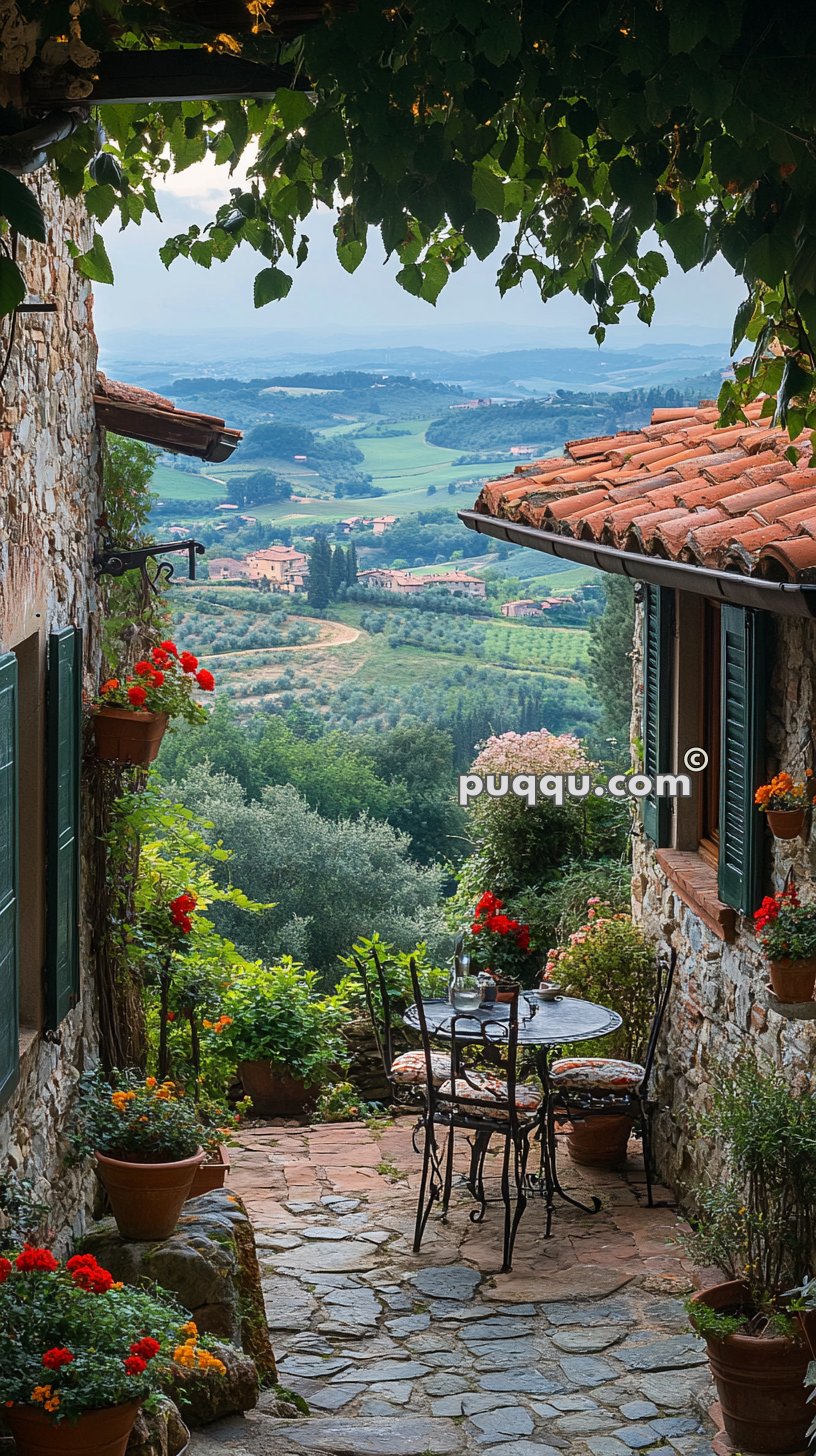 A charming stone patio with potted flowers and an iron table set overlooking a scenic countryside of rolling green hills and scattered houses in the distance.