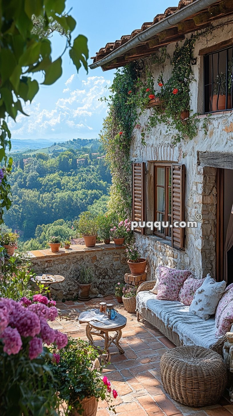 A charming patio with potted plants and flowers, a rustic stone wall with a window featuring wooden shutters, a wicker couch with floral cushions, and a small round table, overlooking a verdant, hilly landscape under a partly cloudy sky.