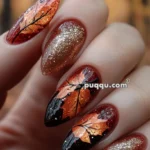 Nails with an autumn-themed design featuring intricate orange leaf patterns and gold glitter accents.