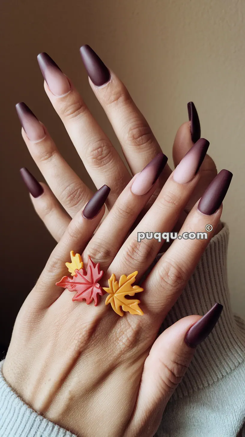 Hands with long, matte, dark burgundy ombre nails, adorned with leaf-shaped rings on the fingers.