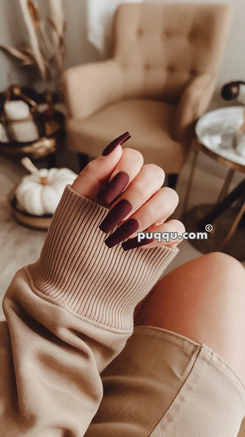 Hand with long, matte burgundy nails held in front of a beige armchair and a living room setting.