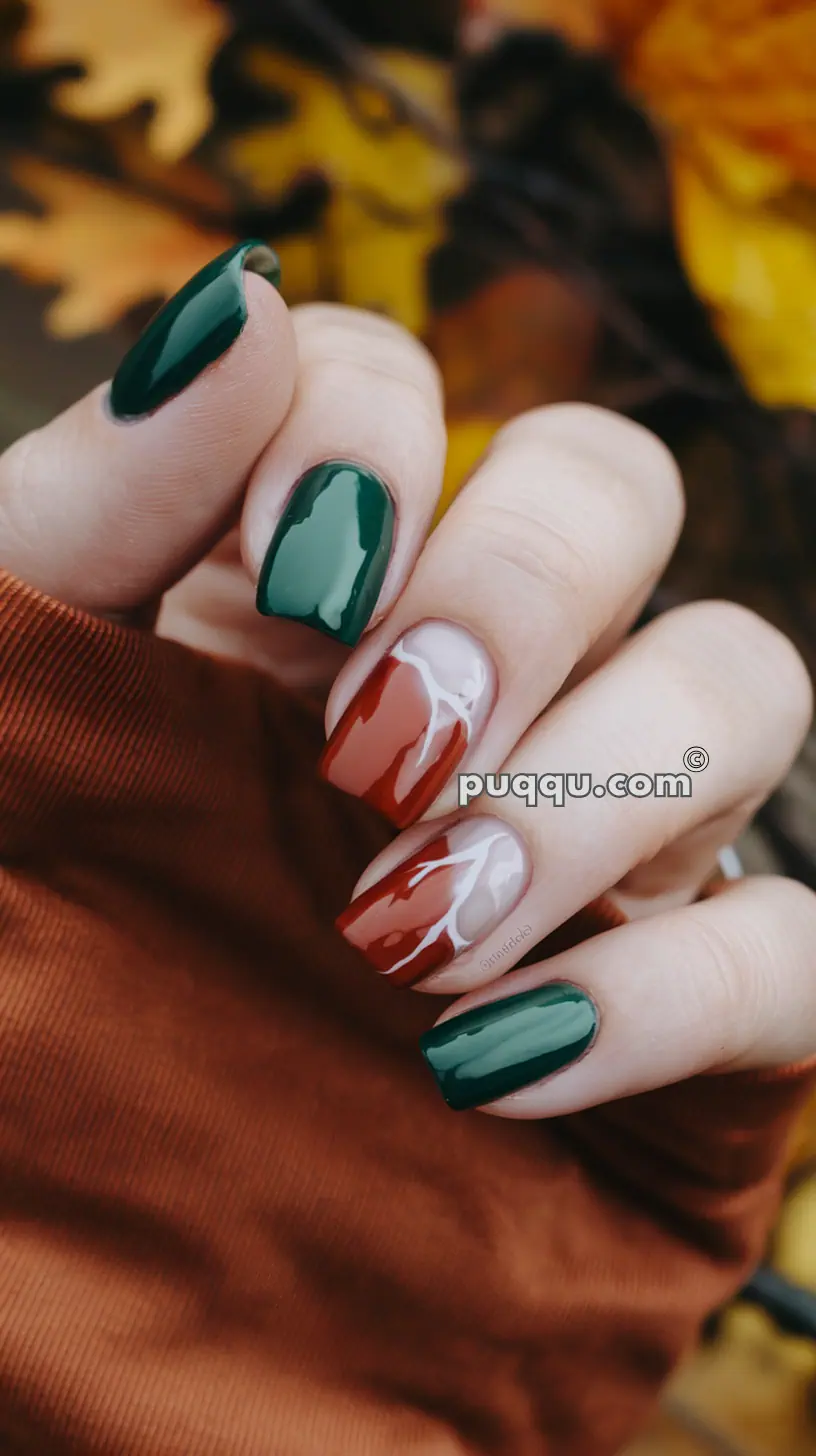 A hand with manicured nails in autumn colors. The nail polish alternates between solid green and red with swirling white accents. The background features blurred fall foliage.