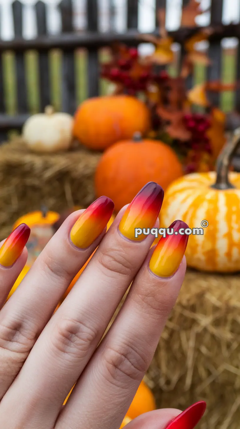 A hand with long, almond-shaped nails featuring a gradient design transitioning from yellow to orange to red, set against a background of pumpkins and fall decorations.