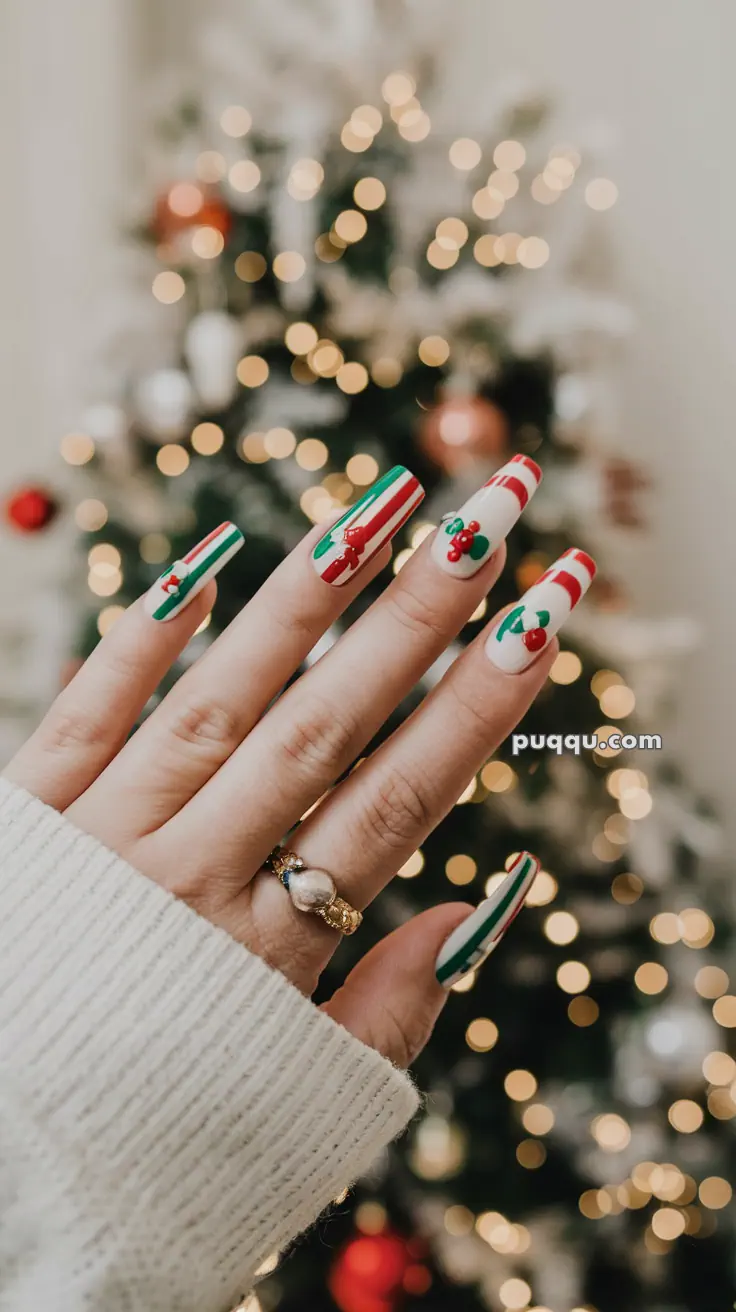 A hand with festive Christmas nail art featuring red, white, and green stripes and holly designs in front of a blurred Christmas tree with lights.