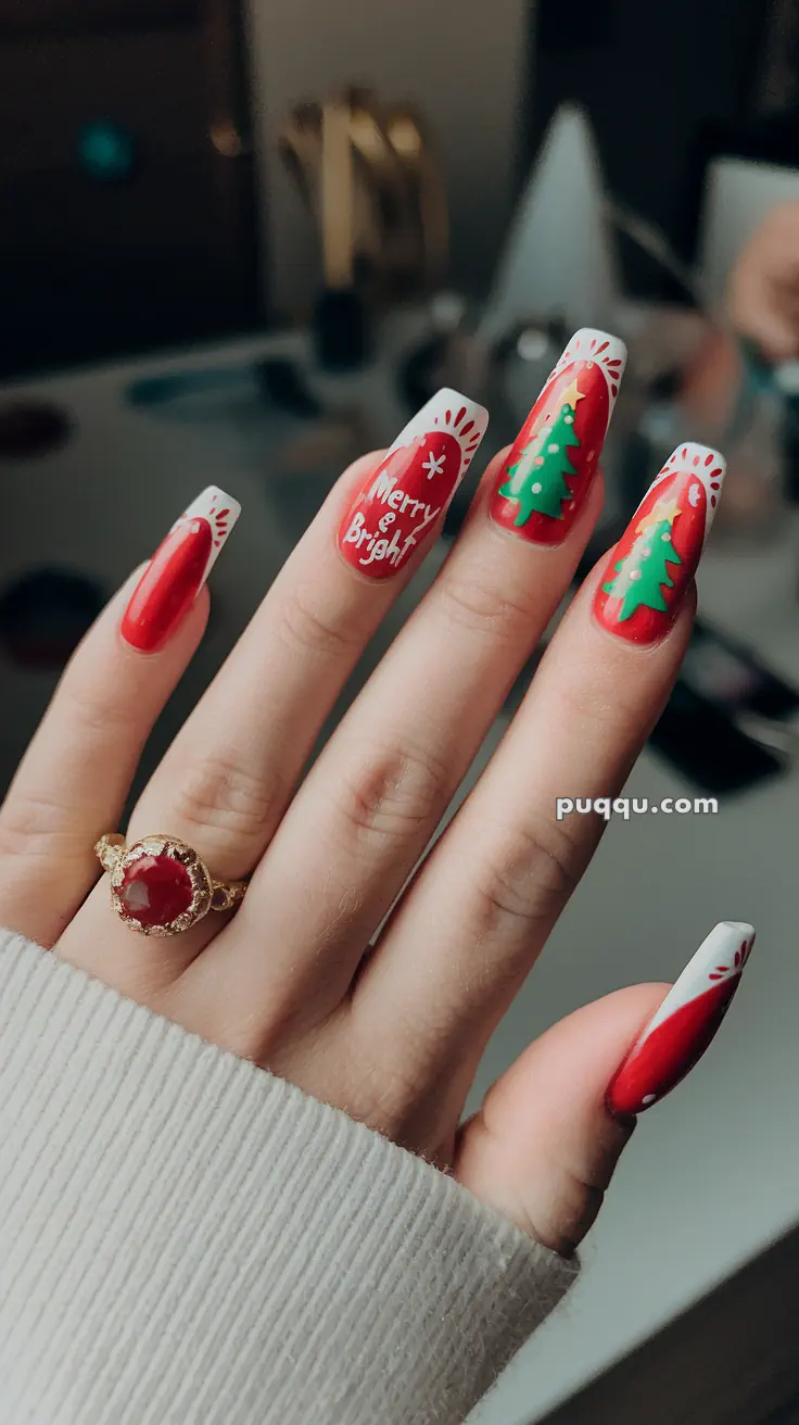 Close-up of a hand with long, festive Christmas-themed nail art featuring red polish, green Christmas trees, and the words "Merry & Bright," adorned with a gold ring with a red stone.