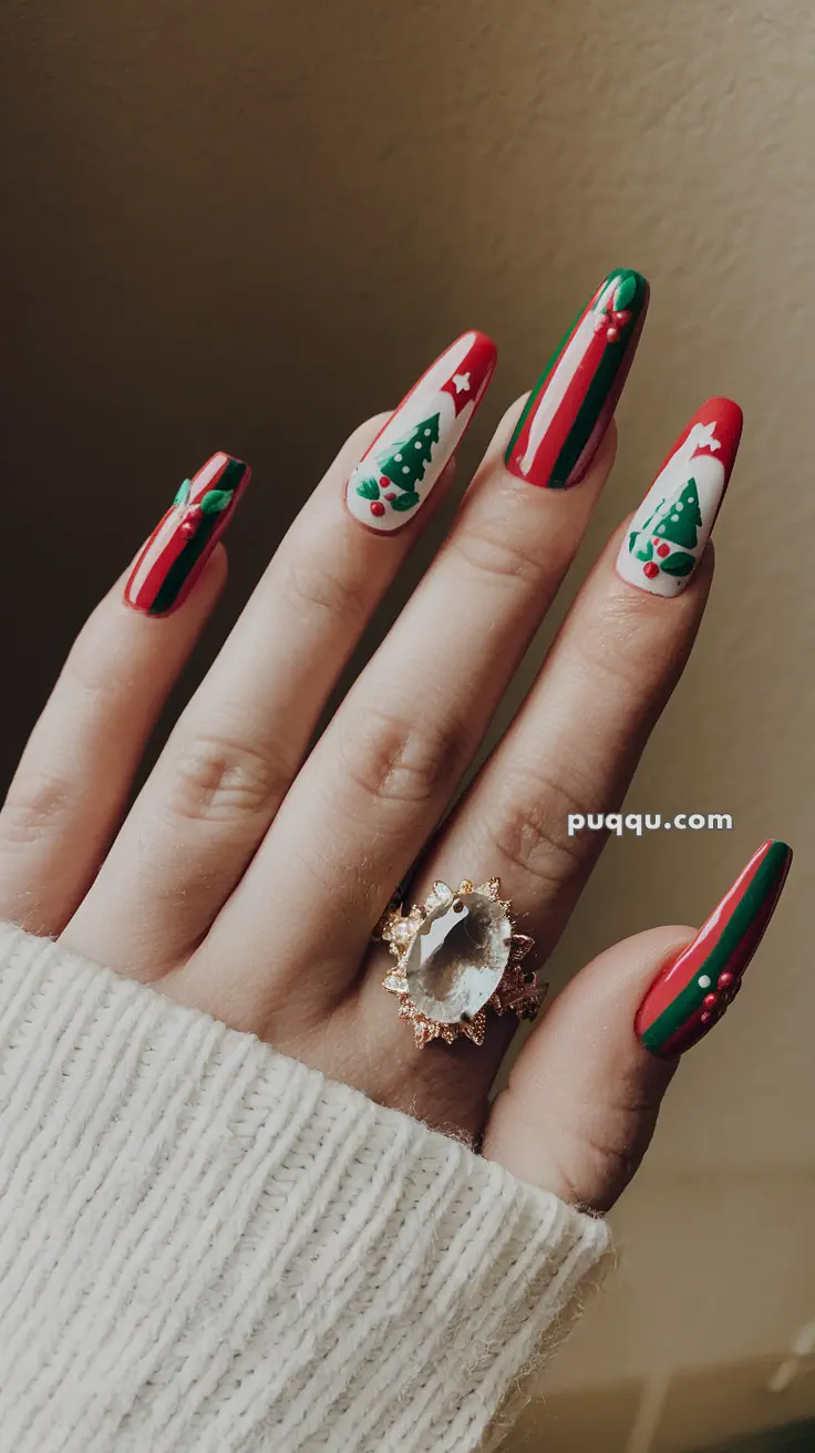 Hand with long nails painted in Christmas-themed designs, featuring green and red stripes, Christmas trees, and holly berries, wearing a large crystal ring.