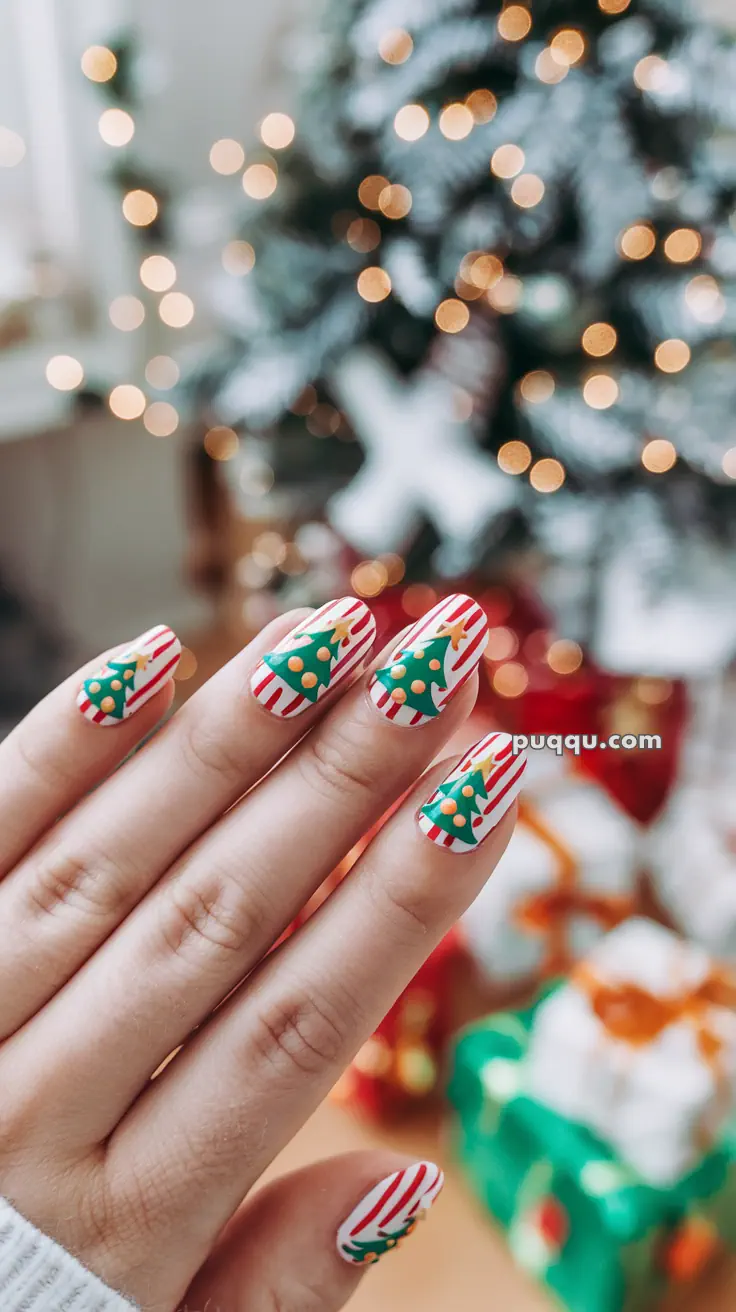 Close-up of a hand with festive Christmas-themed nail art, featuring green Christmas trees on a red and white striped background, against a blurred backdrop of Christmas lights and a decorated tree.