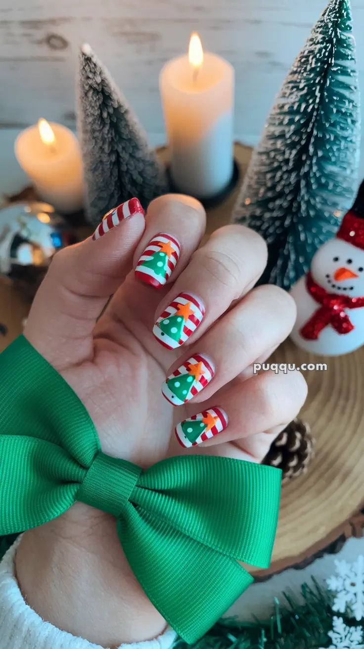 A hand with Christmas-themed nail art featuring red and white stripes, green trees, and stars, holding a green bow, with a festive background including candles, a snowman, and snowy trees.