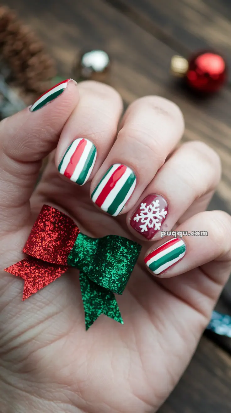 Christmas-themed nail art with red, white, and green stripes, and a white snowflake design, holding a red and green glittery bow.