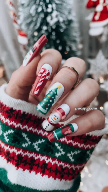 Hand with long festive nails featuring Christmas designs, including a candy cane, Christmas tree, snowman, and gift, with a green, red, and white sweater background.