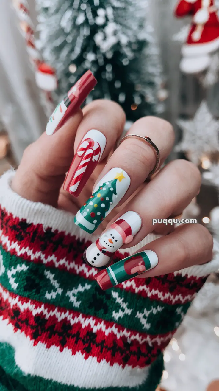 Hand with long festive nails featuring Christmas designs, including a candy cane, Christmas tree, snowman, and gift, with a green, red, and white sweater background.