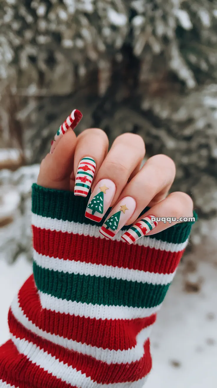 Hand with Christmas-themed nail art, featuring striped designs and Christmas tree patterns, wearing a red, white, and green striped sweater in a snowy outdoor setting.