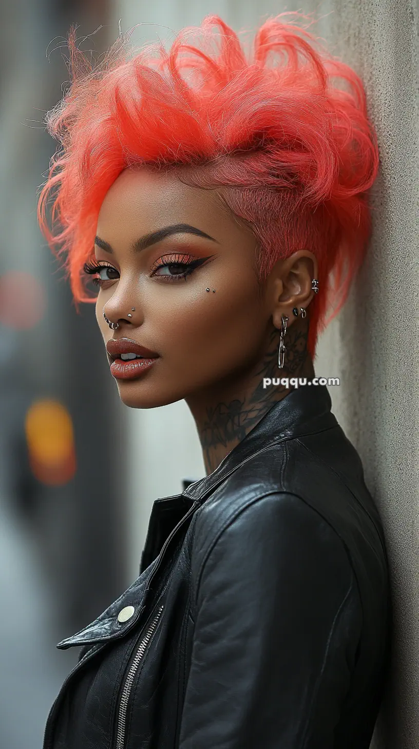 A person with bright orange hair, piercings, and tattoos, wearing a leather jacket, leaning against a wall.