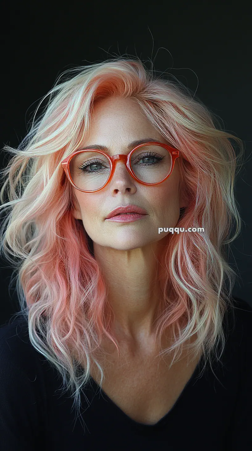 Woman with wavy pink hair and orange glasses against a dark background.