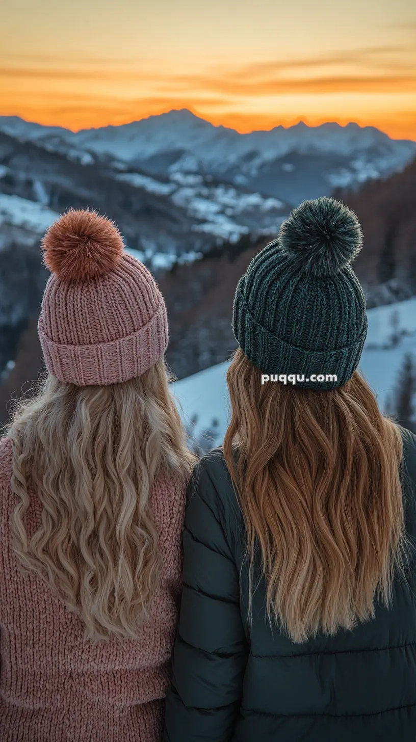 Two people wearing knit hats with pom-poms, standing in front of a snowy mountain landscape during sunset.