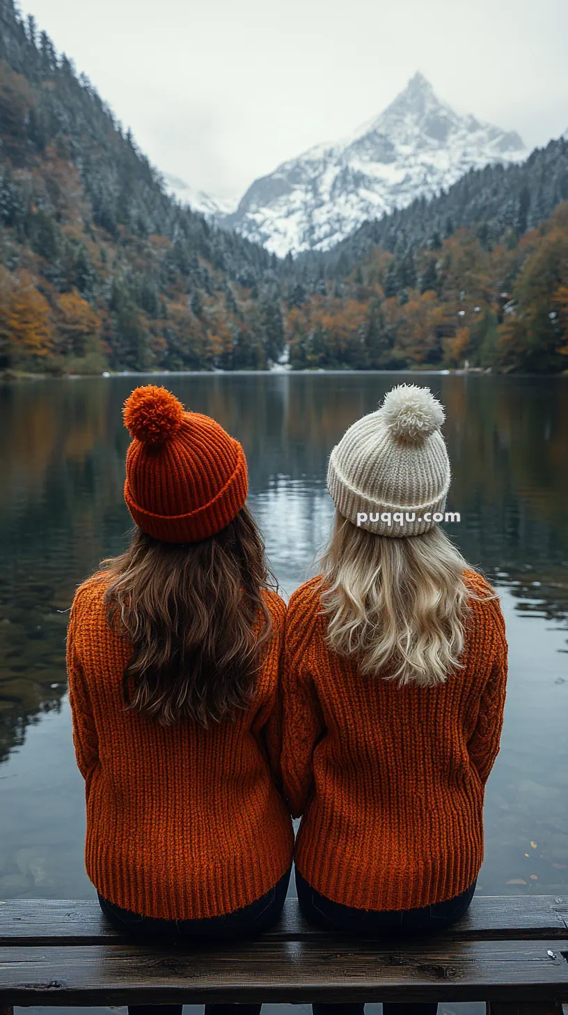 Two people in orange sweaters and knit hats sitting on a bench overlooking a lake with snowy mountains in the background.