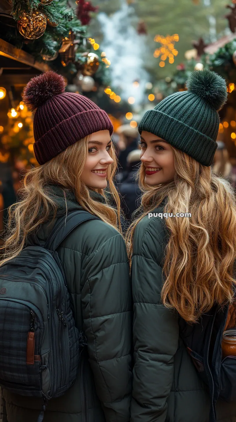 Two people in winter attire with backpacks, wearing knitted hats, smiling in a festive outdoor setting with lights and decorations.