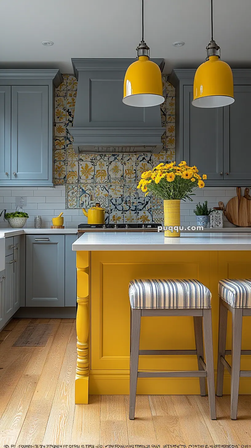 A modern kitchen with blue cabinets, yellow accents including pendant lights and bar stools, a patterned tile backsplash, and a vase with yellow flowers on the island.