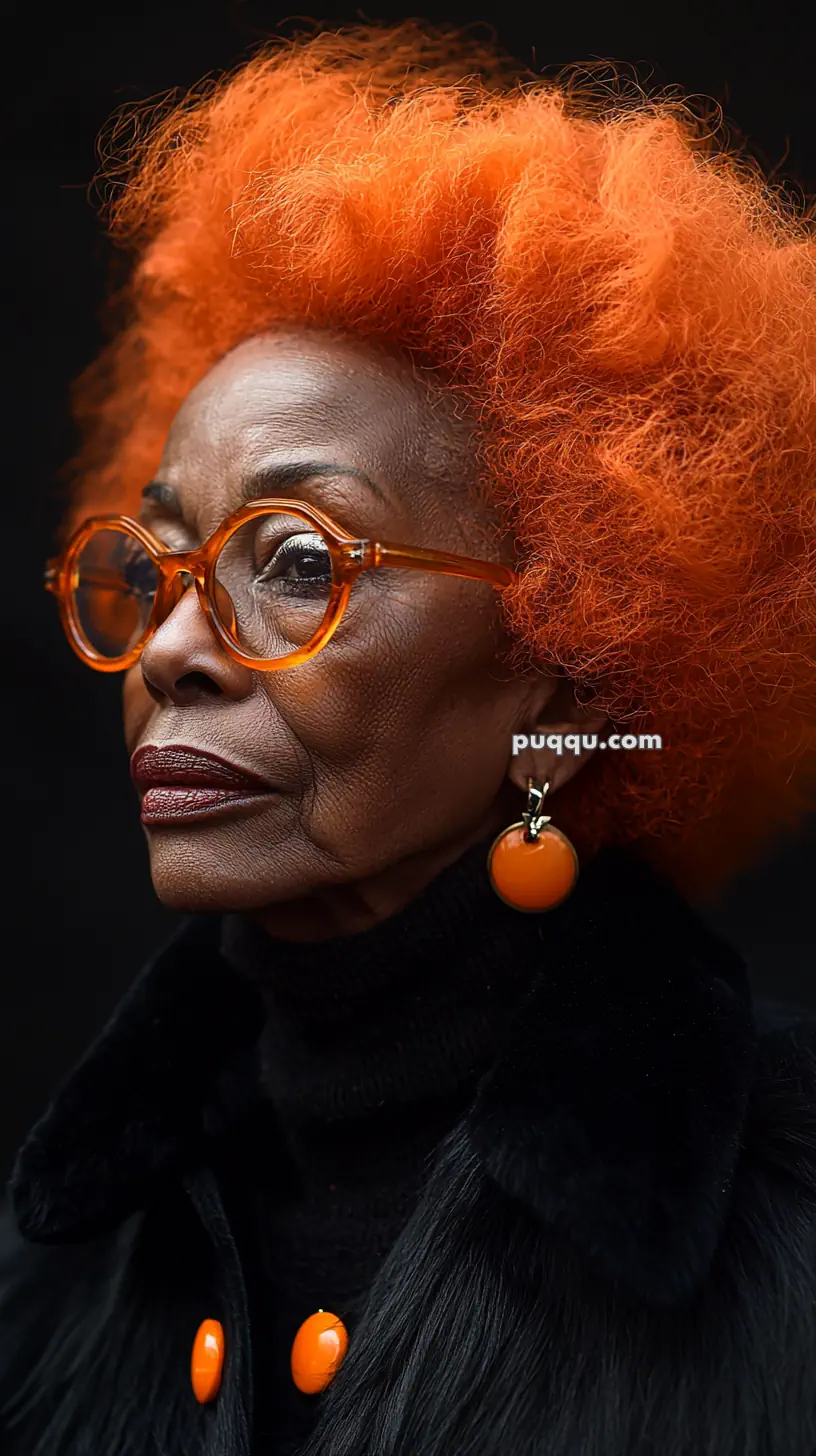 Elderly person with bright orange afro, wearing orange glasses and earrings, in black clothing.