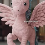 A crocheted pink pegasus toy with blue accents on its hooves and mane, featuring large wings and button-like eyes.
