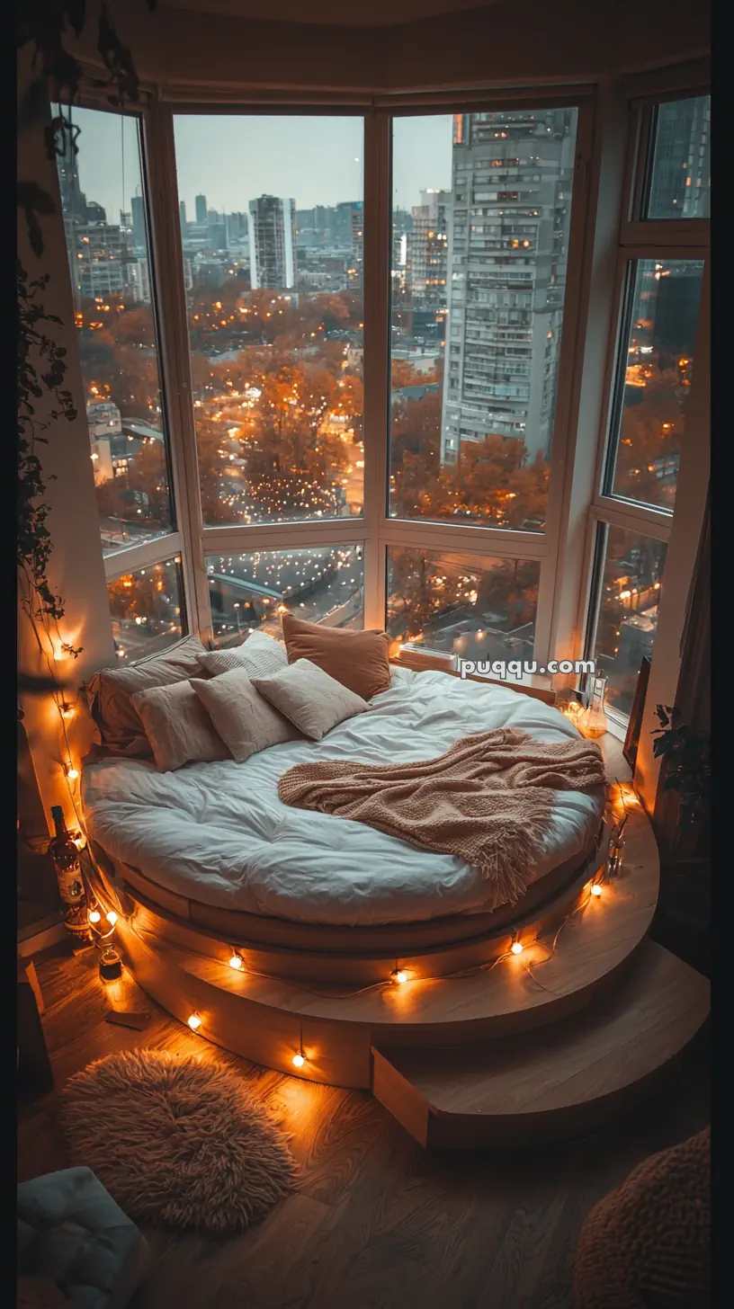 A cozy round bed adorned with white bedding and multiple cushions is placed in front of large, floor-to-ceiling windows offering a view of the city skyline at dusk. The bed is illuminated by warm string lights and surrounded by soft furnishings for a snug atmosphere.