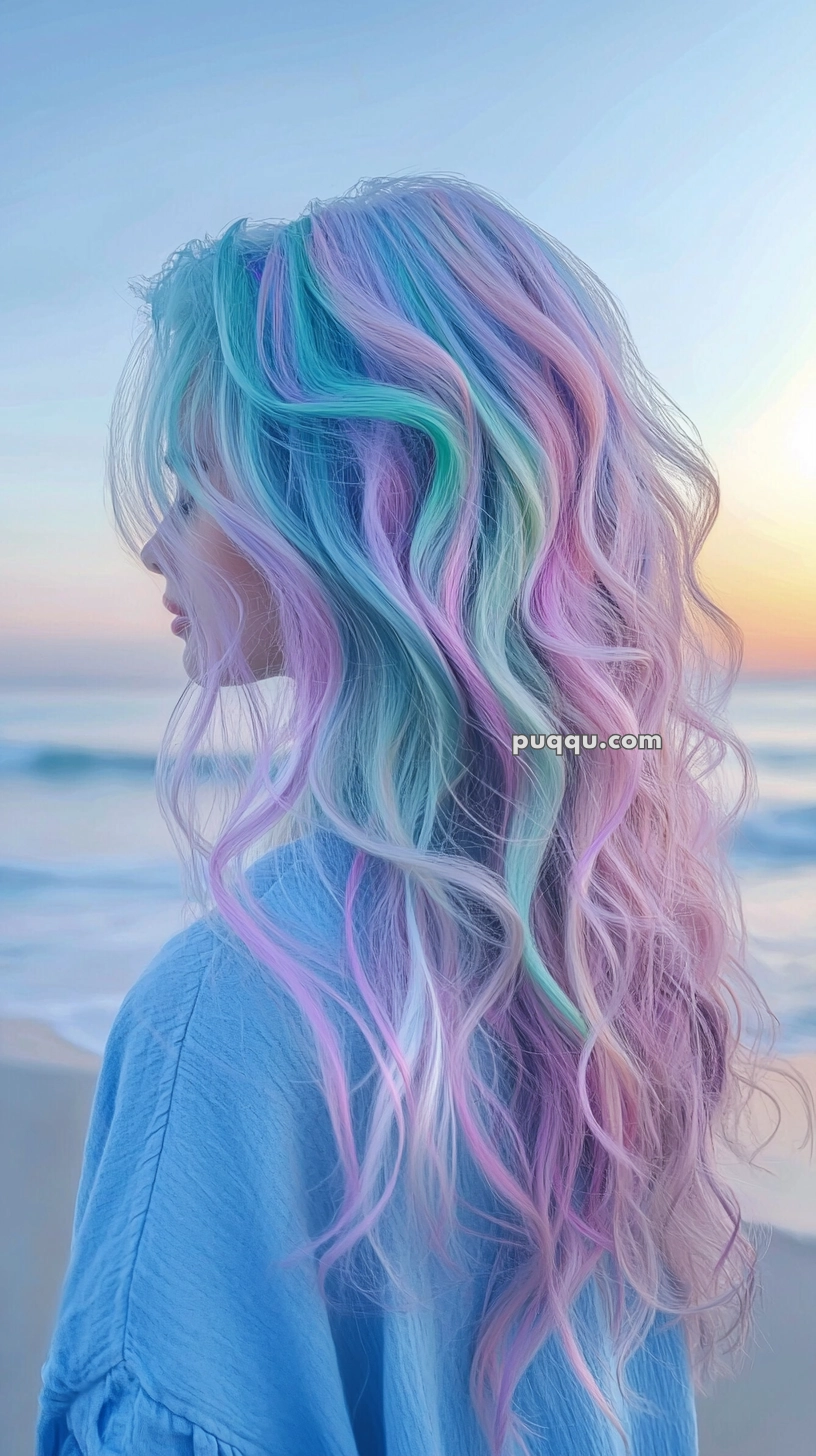 A person with long, wavy hair dyed in pastel colors, including blue, pink, and purple, standing at a beach during sunset.