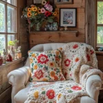A cozy living room corner features a cream-colored armchair with a colorful floral knit blanket and matching pillow. The wooden wall behind is adorned with a shelf holding decorative vases, framed photos, and a bouquet of vibrant flowers. Soft natural light enters through the window, highlighting the rustic decor.