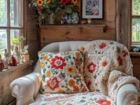 A cozy living room corner features a cream-colored armchair with a colorful floral knit blanket and matching pillow. The wooden wall behind is adorned with a shelf holding decorative vases, framed photos, and a bouquet of vibrant flowers. Soft natural light enters through the window, highlighting the rustic decor.