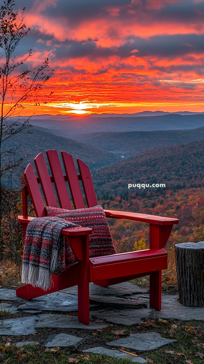 A red Adirondack chair with a colorful plaid blanket draped over it sits on a stone patio overlooking a scenic mountain landscape at sunset. The sky is ablaze with vibrant red, orange, and purple hues, highlighting the distant mountains and forest below.