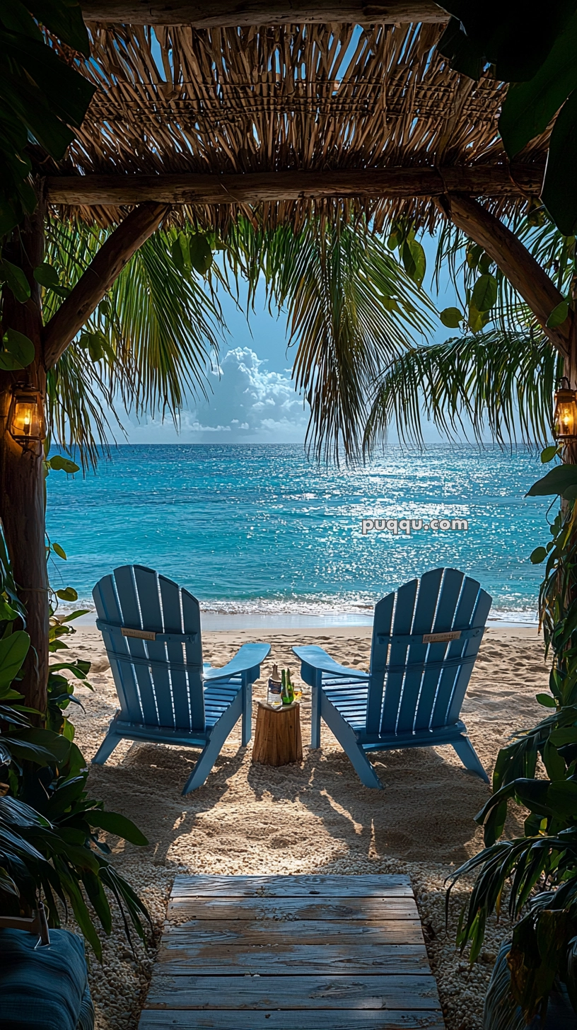 Two blue Adirondack chairs facing the ocean under a wooden pergola, with palm leaves framing the view and a small table holding drinks between them on a sandy beach.