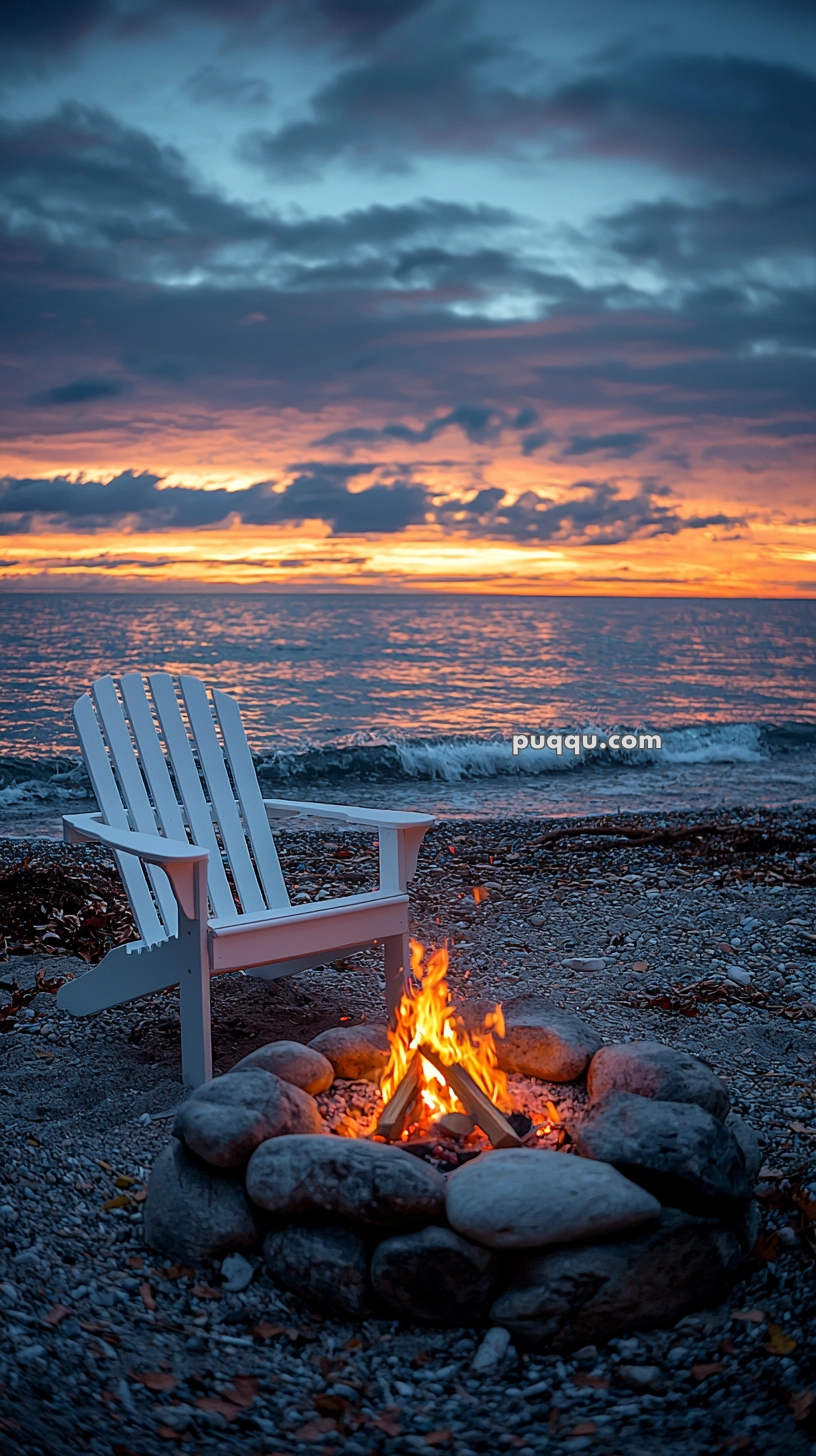 A white Adirondack chair facing a small campfire surrounded by stones on a pebble beach, with a vibrant sunset over the ocean in the background.