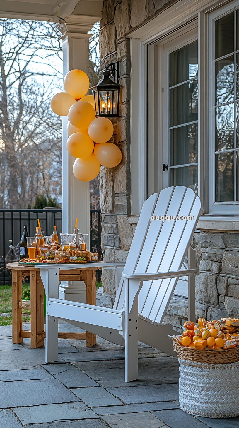 Outdoor patio with a white Adirondack chair, a small wooden table with snacks and drinks, and a basket of oranges. Yellow balloons are attached to a lantern on a stone wall, with trees visible in the background.