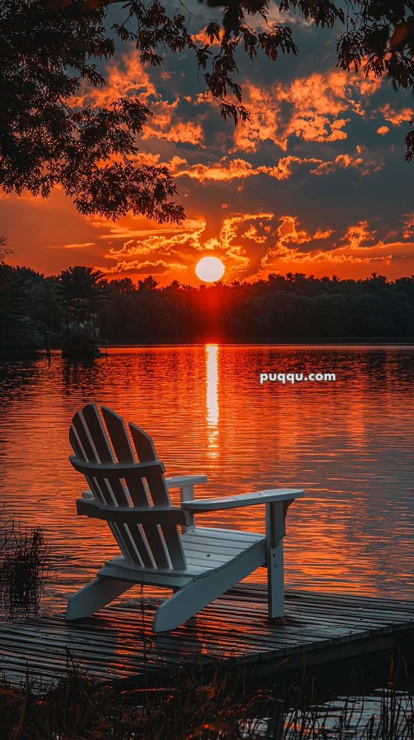 A white Adirondack chair on a wooden dock, facing a serene lake during sunset with the sun casting a vibrant orange glow on the water and clouds.