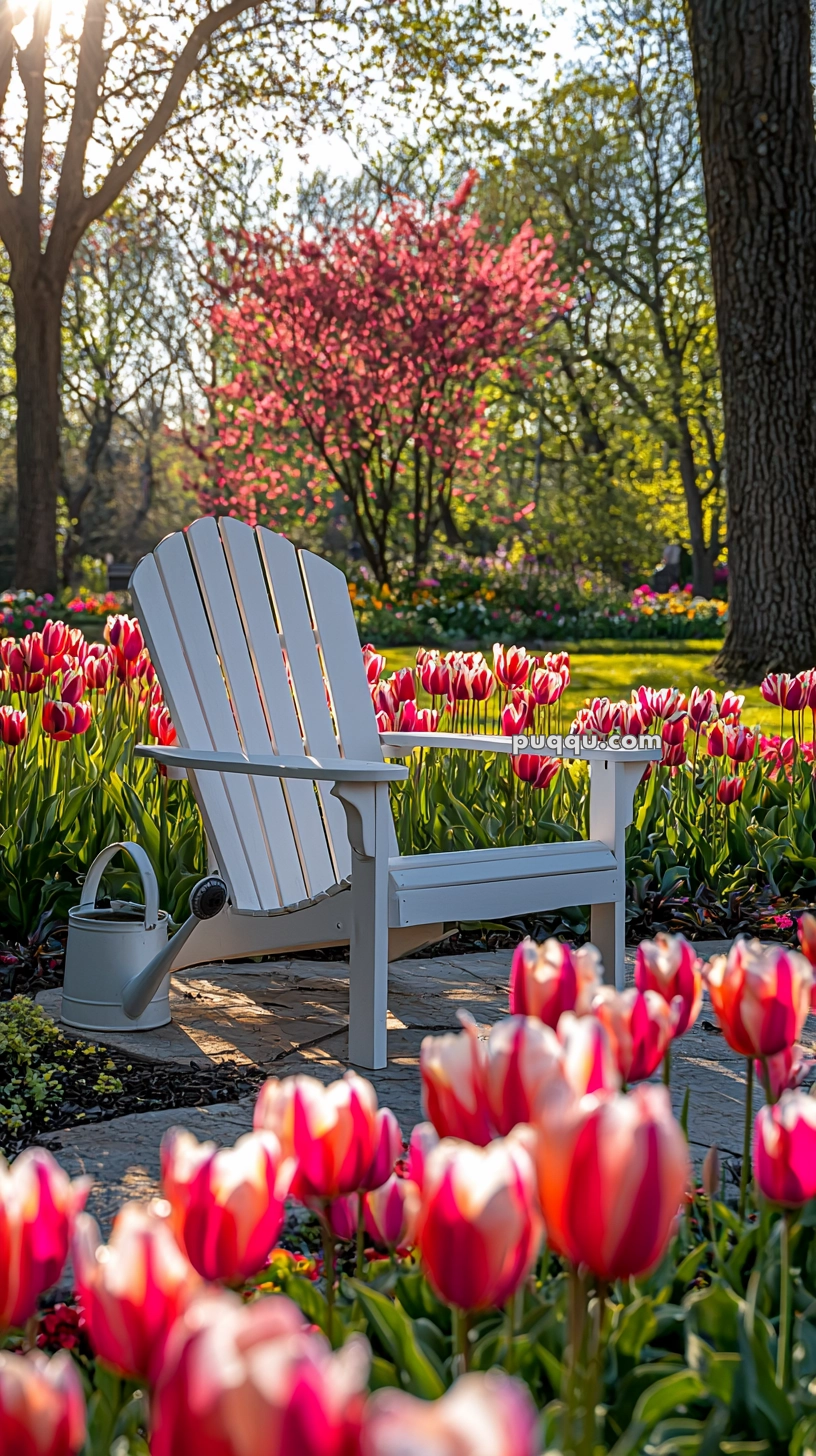 A white Adirondack chair on a stone pathway surrounded by blooming pink and white tulips in a lush garden, with a watering can beside it and pink flowering trees in the background.