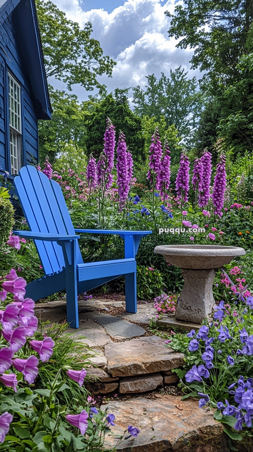 A garden scene with a blue Adirondack chair on a stone patio surrounded by vibrant pink foxgloves and other colorful flowers, next to a small stone birdbath, set against a backdrop of lush greenery and a blue house.