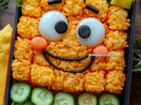A bento box featuring a smiling face made from orange rice, with boiled eggs for eyes, and nori for the mouth and eyebrows. Surrounding the face are fresh lettuce leaves, cucumber slices, and cubes of fruit and vegetables, including yellow and orange pieces. The presentation is colorful and playful.