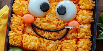 A bento box featuring a smiling face made from orange rice, with boiled eggs for eyes, and nori for the mouth and eyebrows. Surrounding the face are fresh lettuce leaves, cucumber slices, and cubes of fruit and vegetables, including yellow and orange pieces. The presentation is colorful and playful.