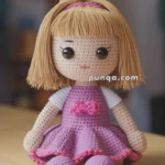 A crocheted doll with blonde hair tied in a bun, wearing a purple dress with a pink bow, and seated on a wooden surface.