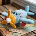 A knitted toy airplane with a blue body, yellow nose, and red wheels, sits atop a world map and a wooden table near a window, with a tag reading "fly high."