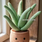 A crocheted potted plant with green leaves sitting on a wooden shelf, featuring a smiling face on the pot.